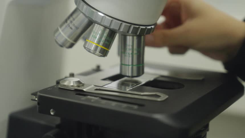 Close-up of a hand changing the objective lens on a microscope to view a slide in a lab