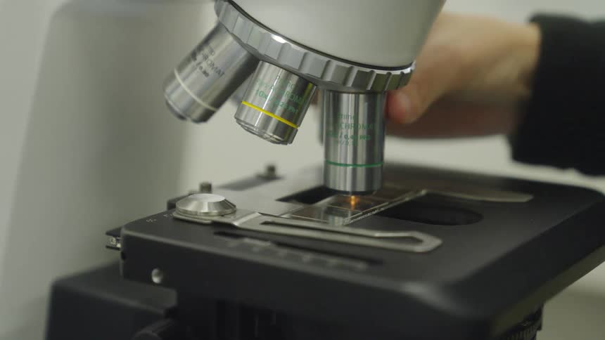 A close-up shot of a scientist's hand adjusting the objective lenses on a modern microscope to examine a specimen slide