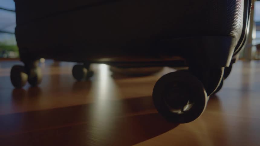 Close-up of a rolling suitcase wheel spinning on a hardwood floor with sunlight creating shadows