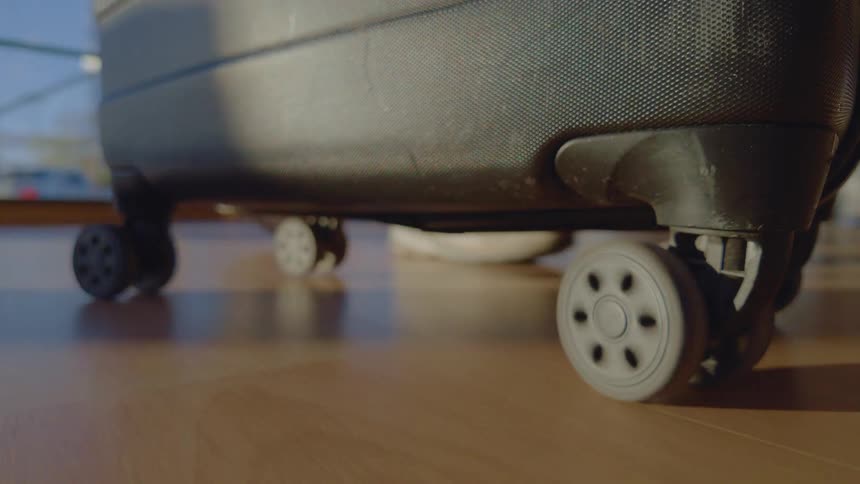 A close-up on the wheels of a black suitcase as it rolls across the sunlit floor of an airport terminal