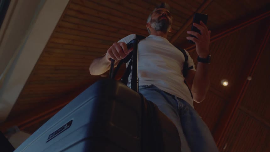 Low-angle perspective of a man with a suitcase checking his phone while standing on a moving walkway at the airport