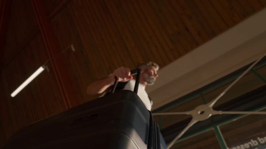 A low angle, slow motion shot follows a rolling suitcase pulled by a mature man walking through a modern terminal