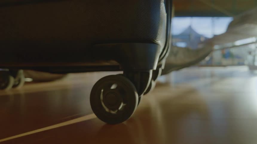 Low-angle close-up of a suitcase wheel rolling across a polished airport floor with sun flares in the background