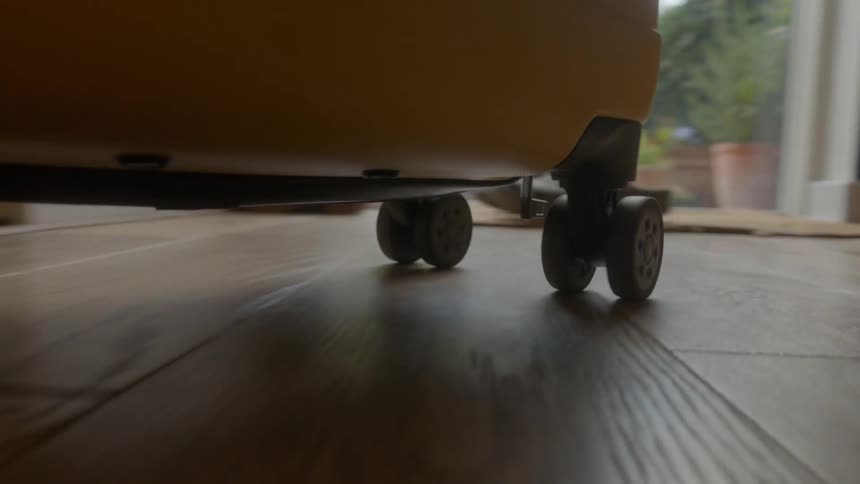 A low-angle close-up of a yellow suitcase's wheels rolling backward across a wooden laminate floor