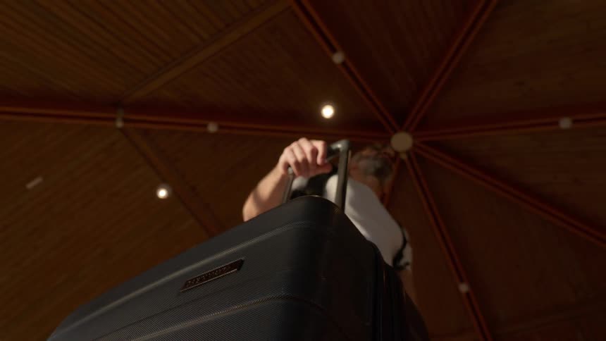 A low-angle, point-of-view shot of a man pulling his rolling luggage through a building with a modern wooden ceiling