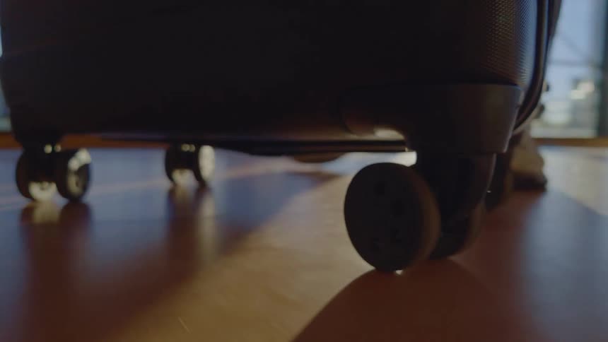 Close-up of a rolling suitcase's wheels spinning across a wooden floor as sunlight casts moving shadows