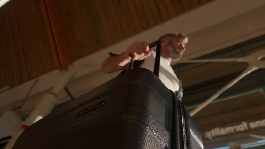 Low angle slow motion shot of a mature man with a beard waiting patiently with his rolling suitcase at a travel terminal