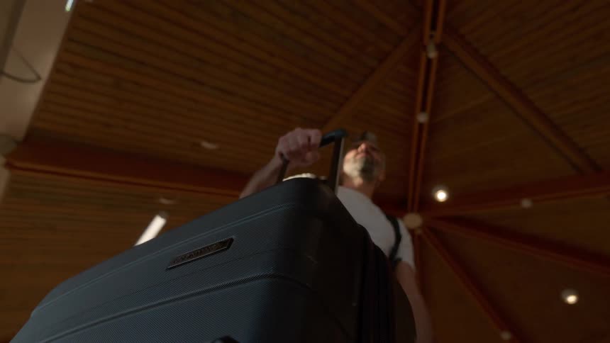 A dramatic low-angle slow-motion shot reveals a man waiting with his rolling suitcase in a modern travel terminal