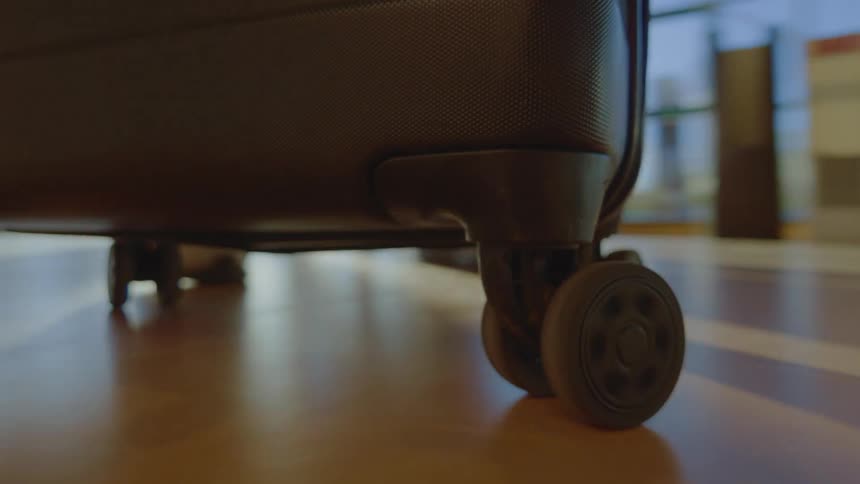 A close-up, low-angle view of a suitcase's wheels rolling across a polished floor in a bright, modern travel terminal