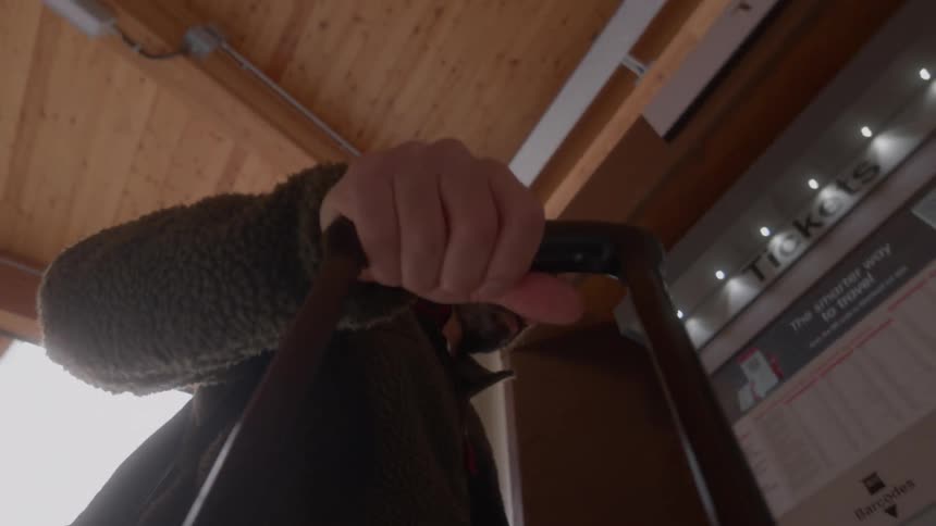 Low angle point-of-view shot of a man pulling a suitcase through a modern train station before stopping at the ticket counter