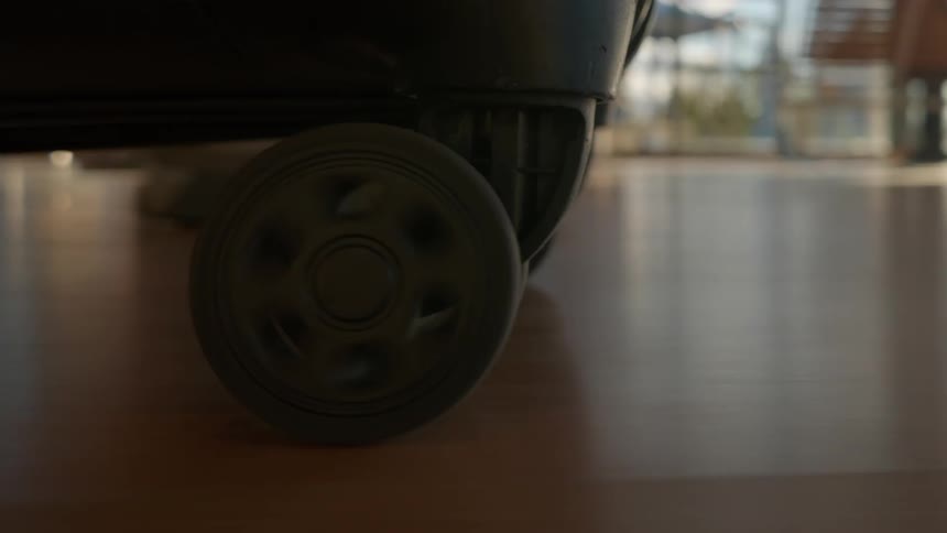 A close-up shot of a suitcase wheel rolling across the polished floor of an airport terminal with a bright sun flare