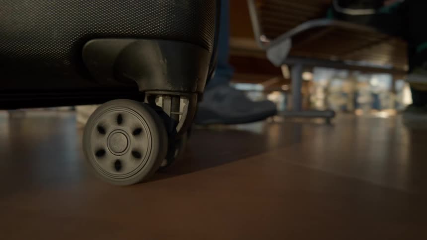 A low-angle close-up follows a spinning suitcase wheel as it rolls across the floor of an airport terminal