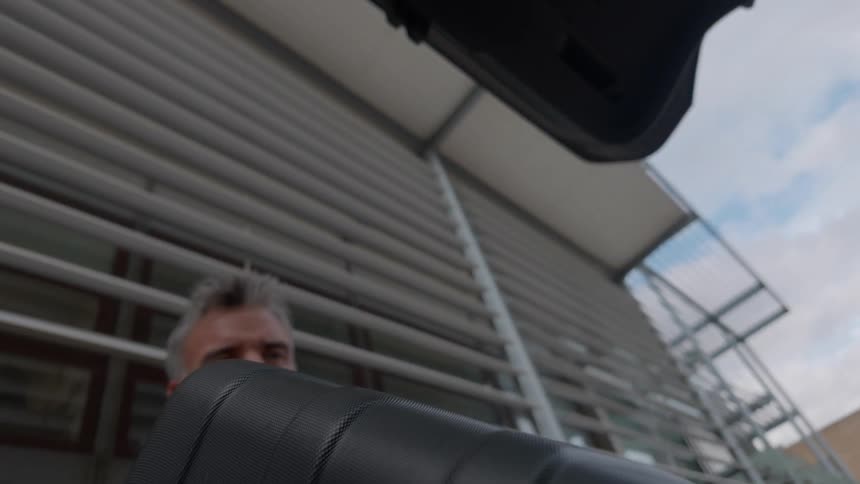 A slow-motion, low-angle shot from inside a car trunk as a man loads a suitcase for an upcoming journey
