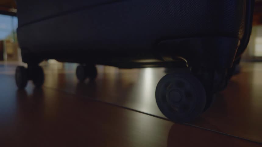 A low-angle close-up of a black travel suitcase's wheels spinning as it is rolled across a shiny floor