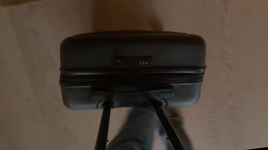 A first-person point of view of a person walking while pulling a black rolling suitcase across a wooden floor