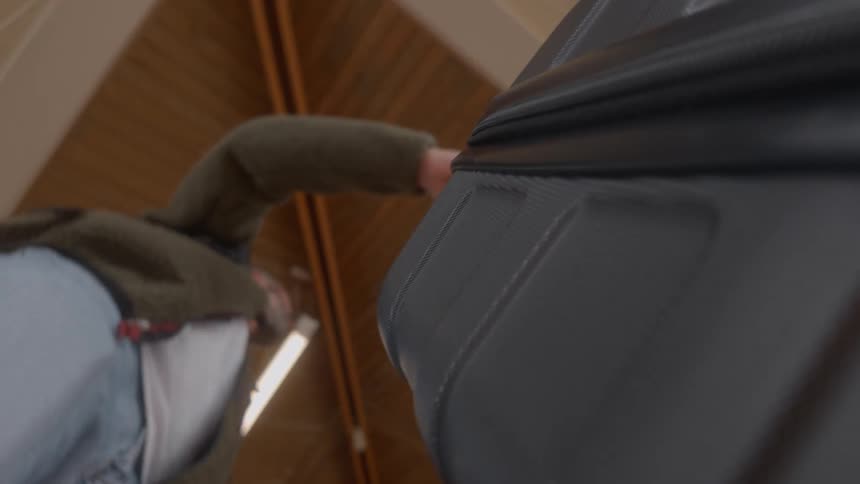 A low-angle point-of-view shot shows a man struggling to zip up an overstuffed suitcase