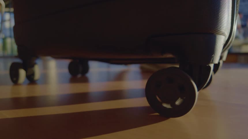 A close-up shot of the wheels of a black travel suitcase on a shiny floor as light and shadows move across it