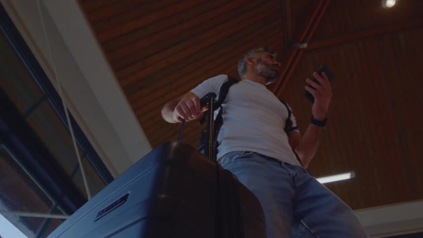 A low-angle view shows a male traveler with a suitcase and smartphone waiting inside a modern airport terminal with a high wooden ceiling