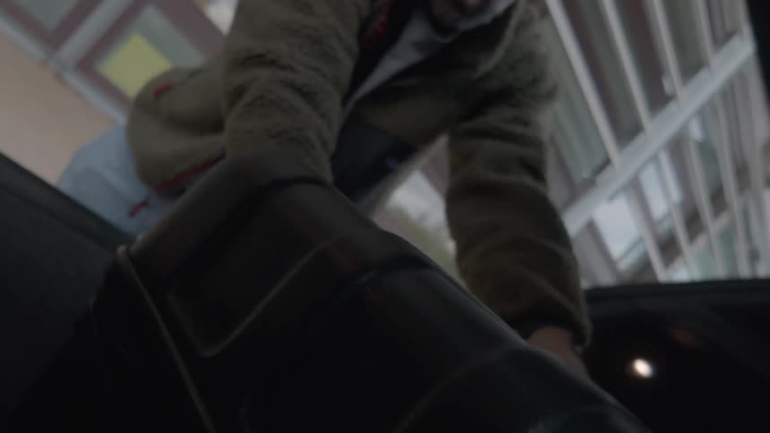 A man unloads his black hardshell suitcase from the trunk of a car, filmed from a low angle inside the vehicle