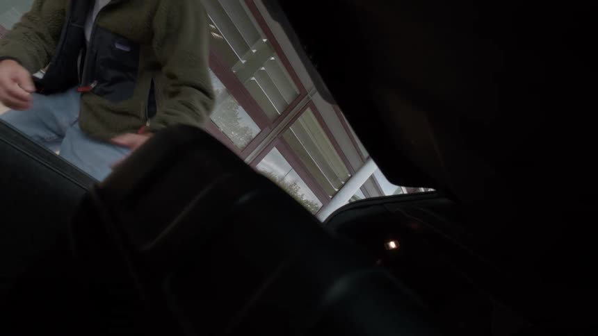 Shot from inside a car trunk, a man opens it remotely and pulls out a rolling suitcase before walking towards an airport