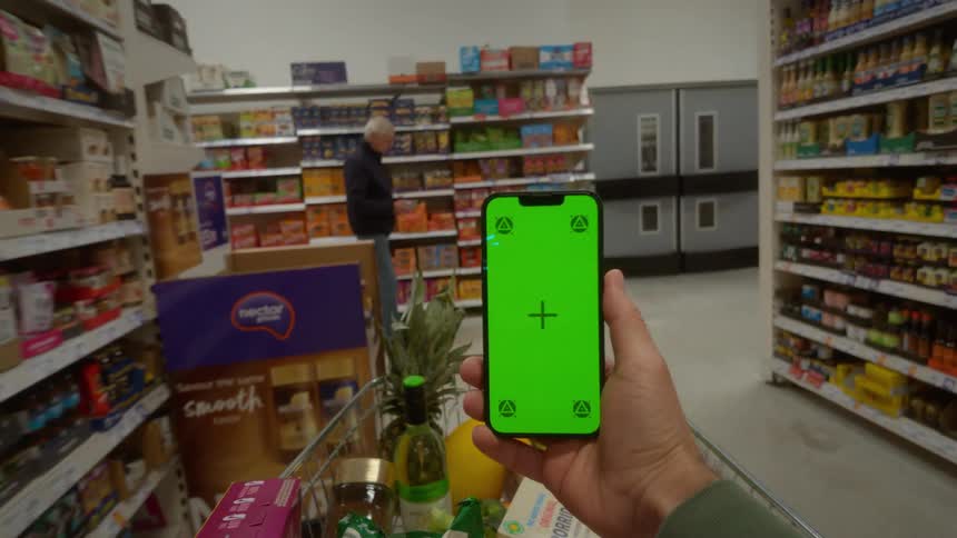 A first-person view of a shopper holding a smartphone with a green screen while pushing a cart down a grocery store aisle