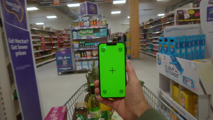 A first-person view of someone pushing a shopping cart through a grocery store while holding a smartphone with a green screen
