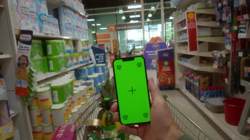 First-person perspective of a shopper pushing a cart down a supermarket aisle while holding a smartphone with a green screen