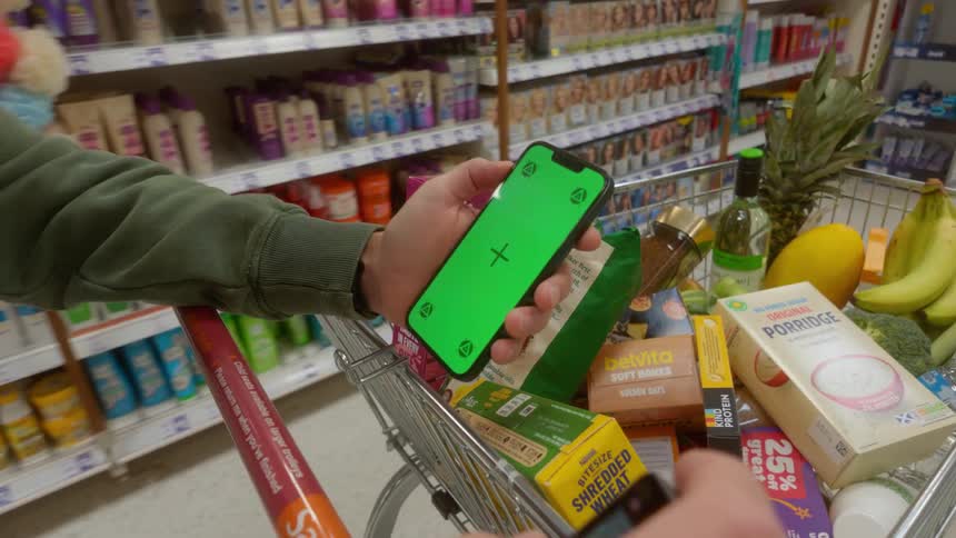 Shopper's perspective holding a green screen smartphone over a full shopping cart in a grocery store