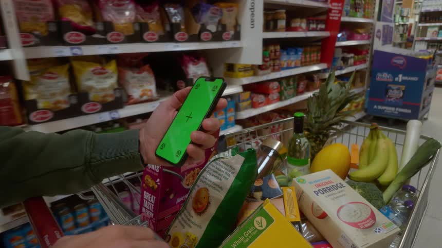 A first-person point of view of a man holding a green screen smartphone while pushing a full shopping cart down a grocery store aisle