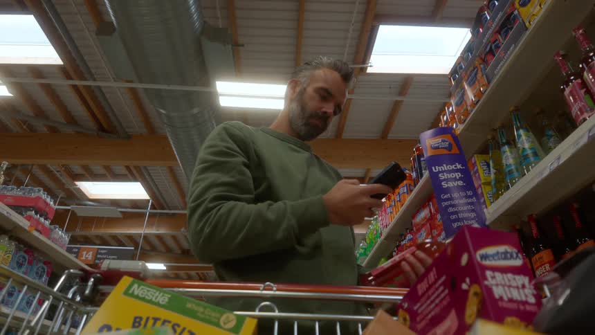 A man in a supermarket scans a bottle with a self-scan device, shot from a low angle inside the shopping cart
