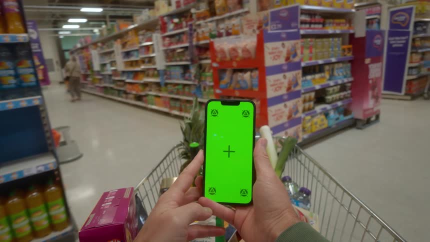 A first-person point of view shot of a person using a green screen smartphone while pushing a shopping cart through a supermarket aisle