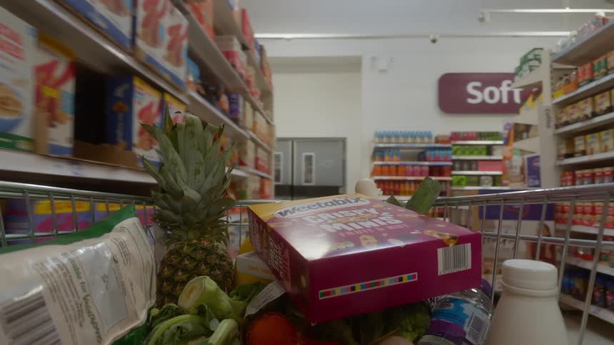 A slow-motion point-of-view shot from inside a shopping cart as a box of cereal is added before moving down the grocery store aisle