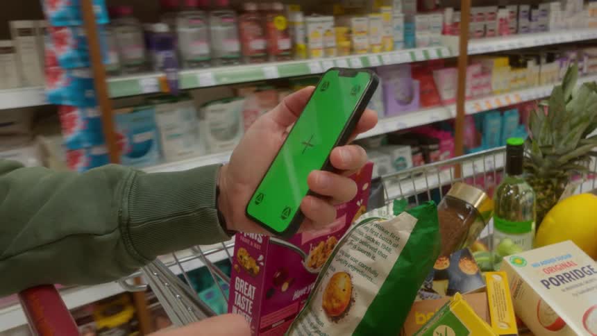 A person's hand holds a smartphone with a green screen while pushing a shopping cart through the aisles of a grocery store