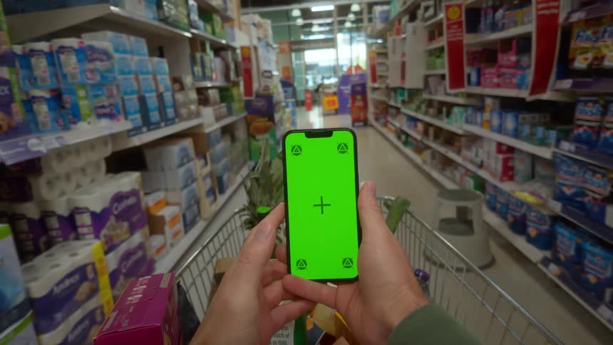 A first-person perspective of a shopper pushing a cart down a supermarket aisle while holding a smartphone with a green screen