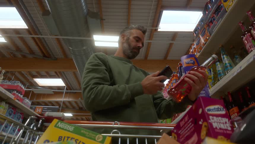 From a low angle inside a shopping cart, a man uses a self-scanner on a drink in a supermarket aisle