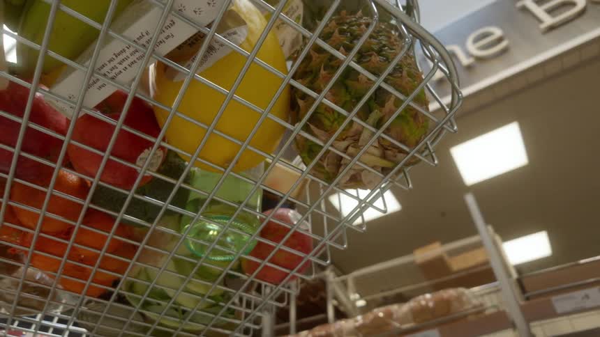 A low-angle point-of-view shot from inside a wire shopping basket filled with fresh fruit as it moves through a supermarket bakery