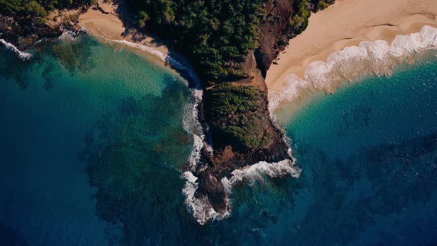 A stunning aerial top-down view of a lush green rocky peninsula separating two pristine sandy beaches with turquoise and deep blue ocean waves