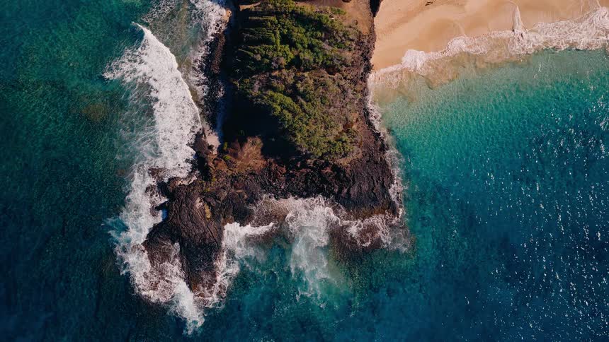 An immersive top-down aerial cinemagraph of a tropical volcanic headland where vibrant turquoise ocean waves crash against the rocky shore and a golden sand beach