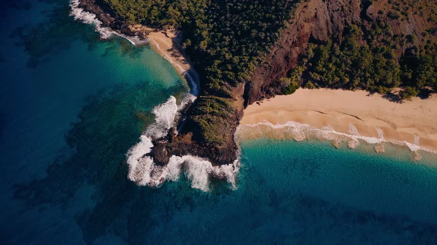 A static top-down aerial view shows gentle waves breaking on a rocky headland separating two pristine sandy beaches with turquoise water