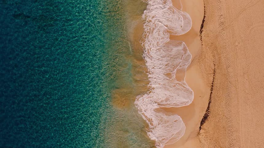 An aerial top-down slow-motion view shows tranquil turquoise waves gently washing over a golden sandy beach
