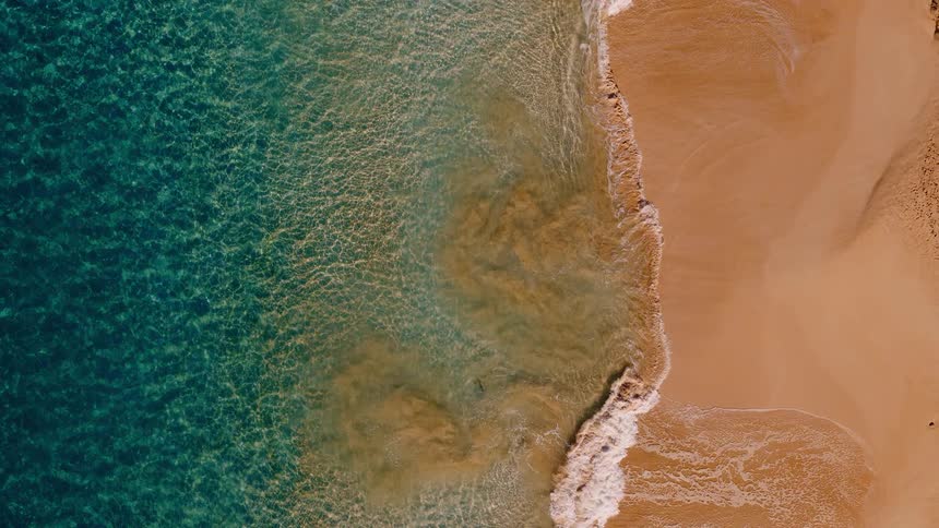 An aerial top-down view shows vibrant turquoise ocean water meeting a golden sandy beach as gentle waves roll ashore