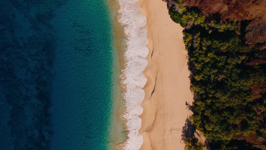 A mesmerizing top-down aerial view shows gentle turquoise waves washing over a secluded golden sand beach