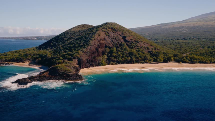 An aerial view pulls back from a secluded tropical beach with turquoise water nestled at the base of a volcanic hill