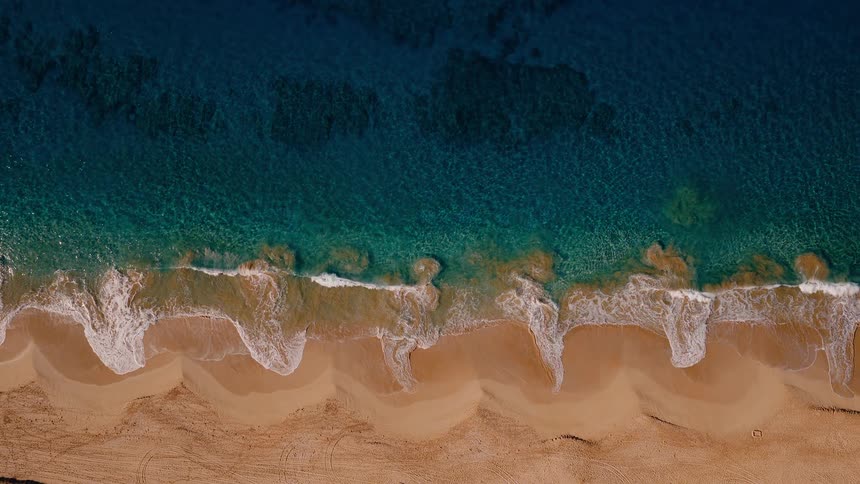 A mesmerizing top-down aerial view of clear turquoise ocean waves slowly breaking on a beautiful sandy beach