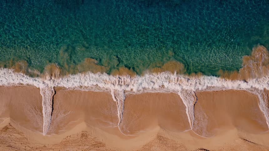 A beautiful top-down aerial shot captures turquoise ocean waves gently rolling and breaking onto a golden sandy beach in slow motion