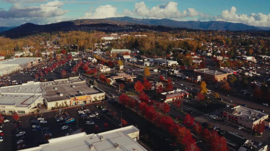 An aerial drone view slowly pushes in on a vibrant small town with red autumn foliage, a shopping center, and distant mountains