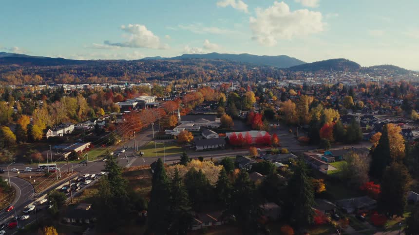 An aerial drone shot slowly rises to reveal a quaint suburban town bathed in the warm light of a sunny autumn day, showcasing vibrant fall foliage and distant hills