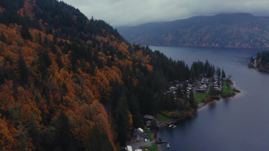 A slow ascending aerial shot captures a tranquil lakeside community surrounded by mountains covered in vibrant autumn foliage on a cloudy day