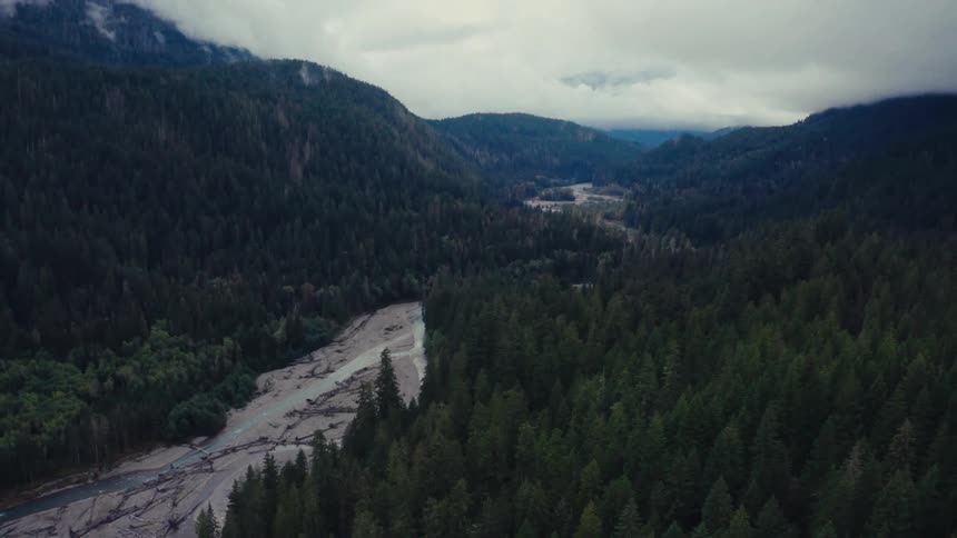A slow aerial pullback reveals a winding river cutting through a vast, moody evergreen forest valley under a cloudy sky