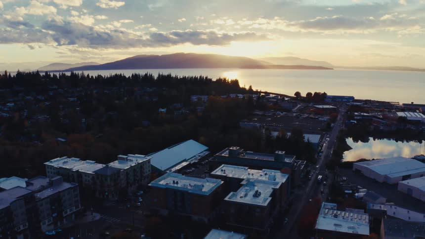 An aerial shot flies over a small coastal town at dusk, tilting up to reveal a beautiful sunset over the water and distant islands